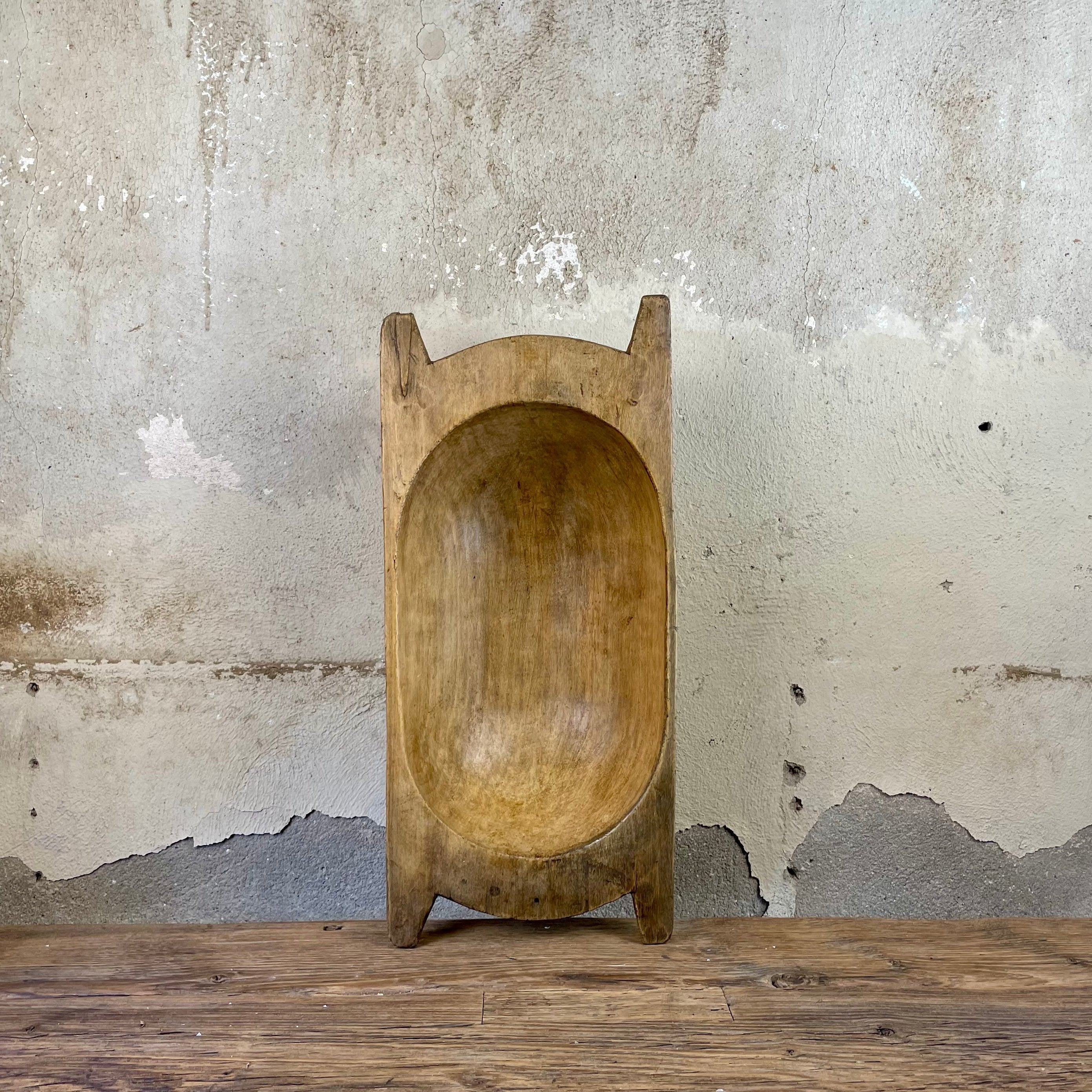 Oval hand-carved wooden dough bowl with handle ends, standing upright against a plaster wall