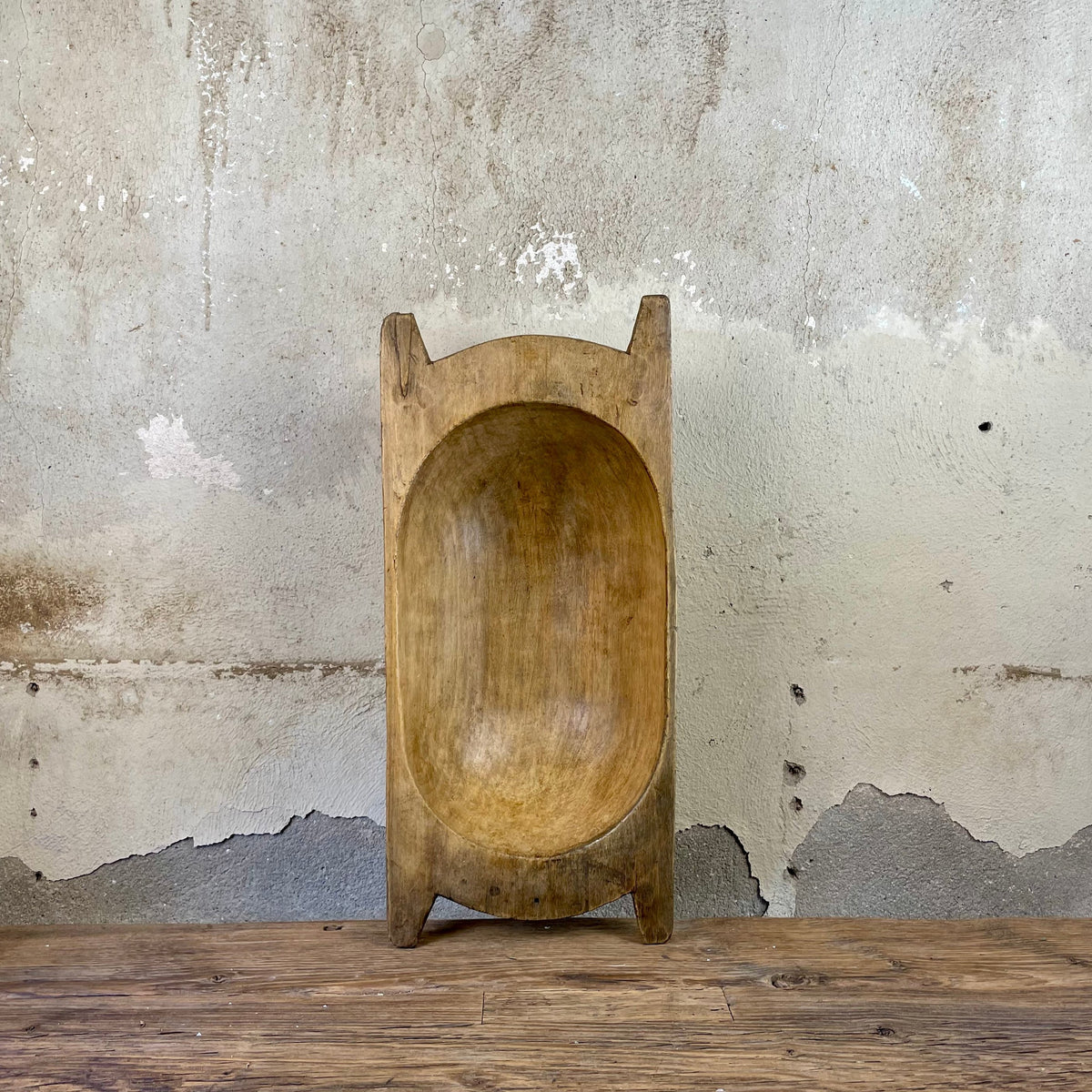 Oval hand-carved wooden dough bowl with handle ends, standing upright against a plaster wall