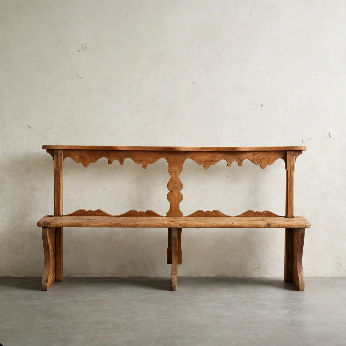 A rustic wooden bench with a scalloped apron and turned central post, shown against a plain light gray wall and concrete floor.